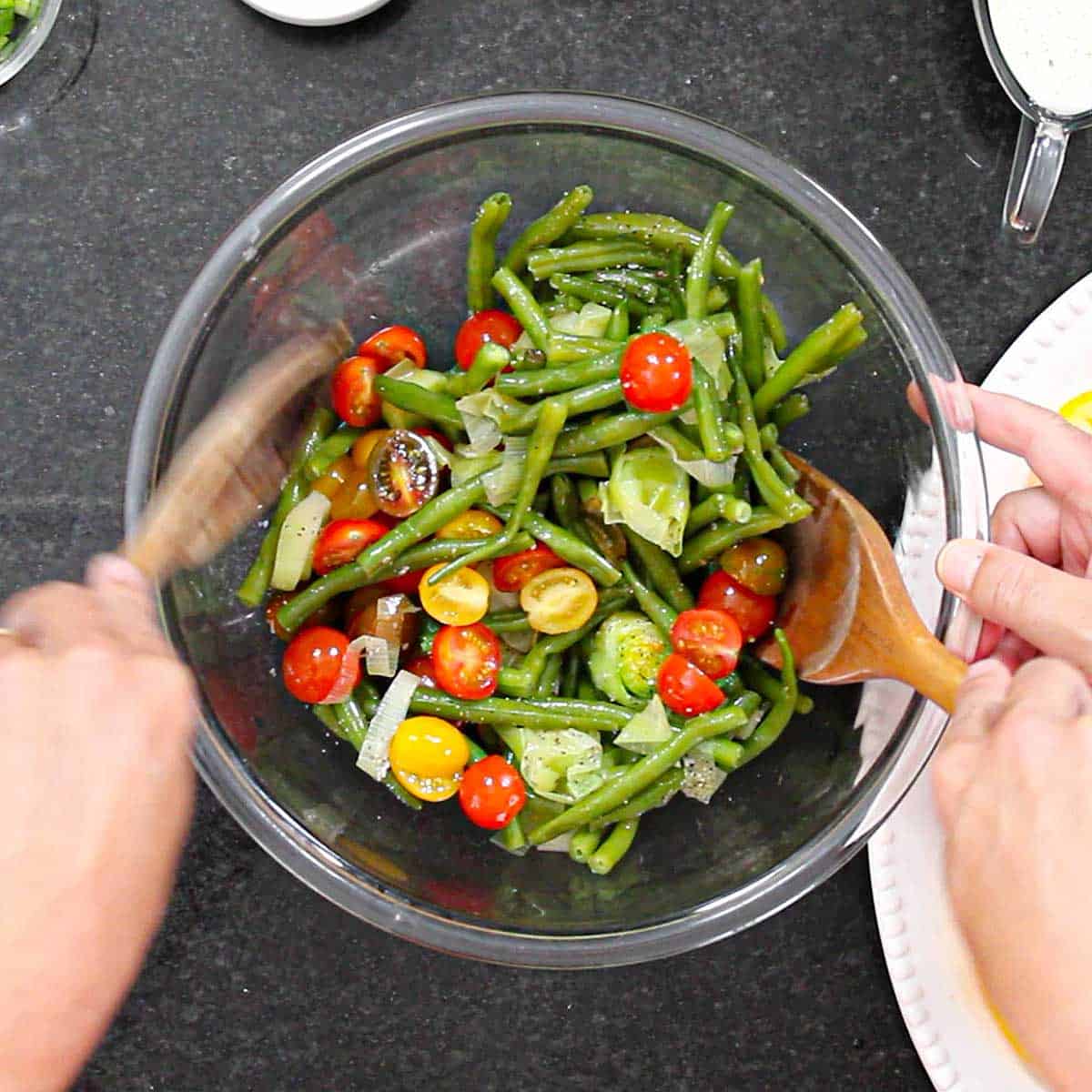 A person using two wooden spoons to toss together blanched green beans and slice leeks with halved cherry tomatoes, salt, and pepper.