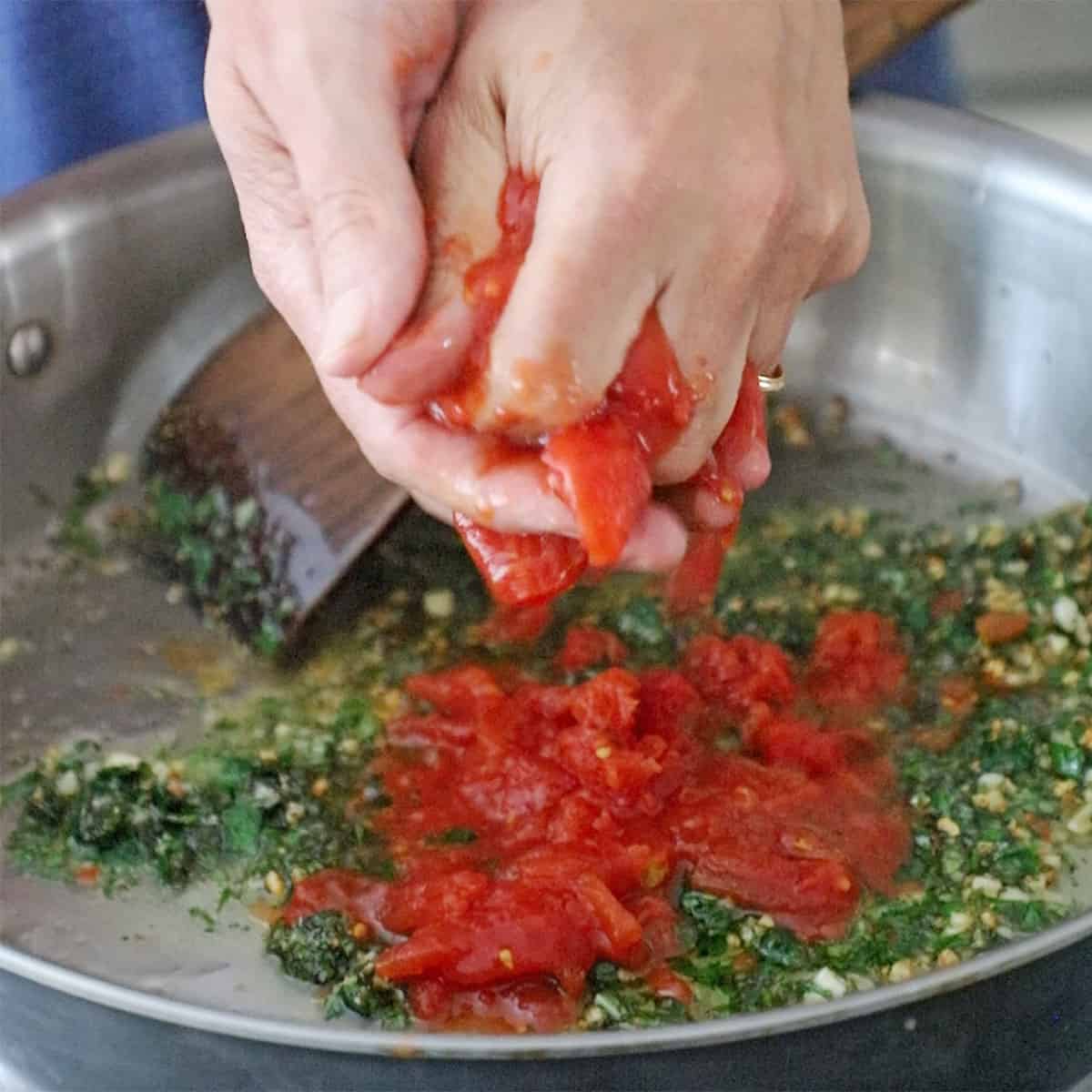 A person using both of their hands to squeeze whole tomatoes into a large skillet filled with sautéed herbs and garlic in olive oil.