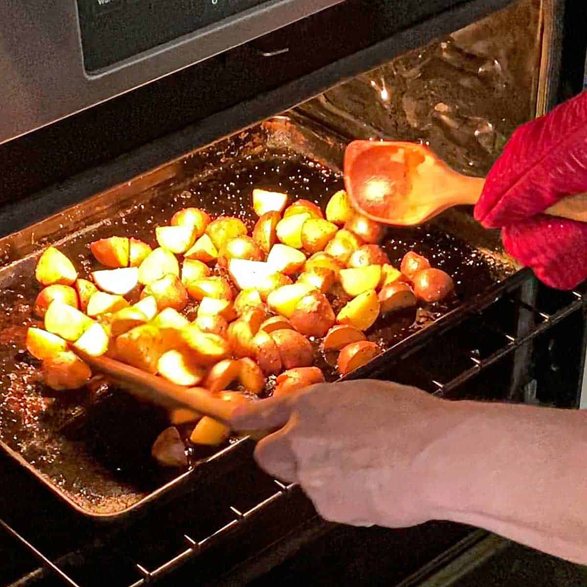 A person holding wooden spoons in both hands and using them to toss new potatoes that are roasting on a pan inside an oven.