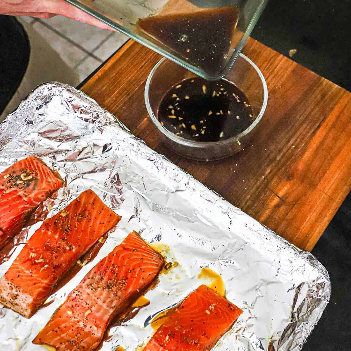 A person pouring teriyaki salmon marinade from a glass baking dish into a small glass bowl next to four uncooked marinated salmon fillets on a foil-lined baking sheet.