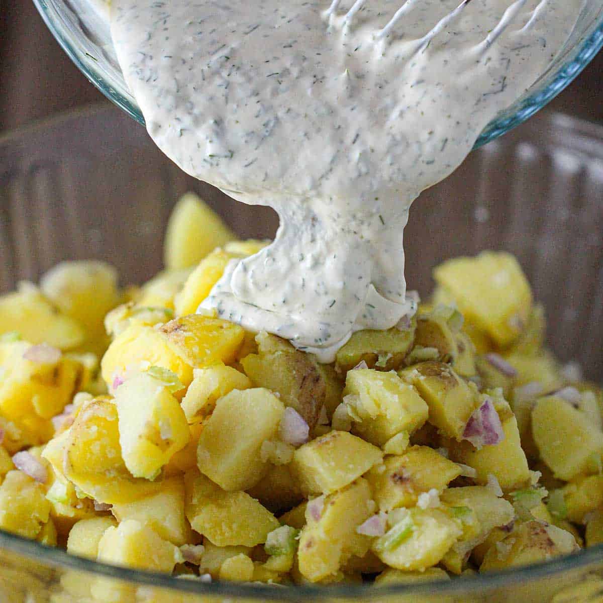 A person pouring freshly prepared dill and mayonnaise dressing over cooked potatoes that have been tossed with chopped red onion and celery in a glass bowl.