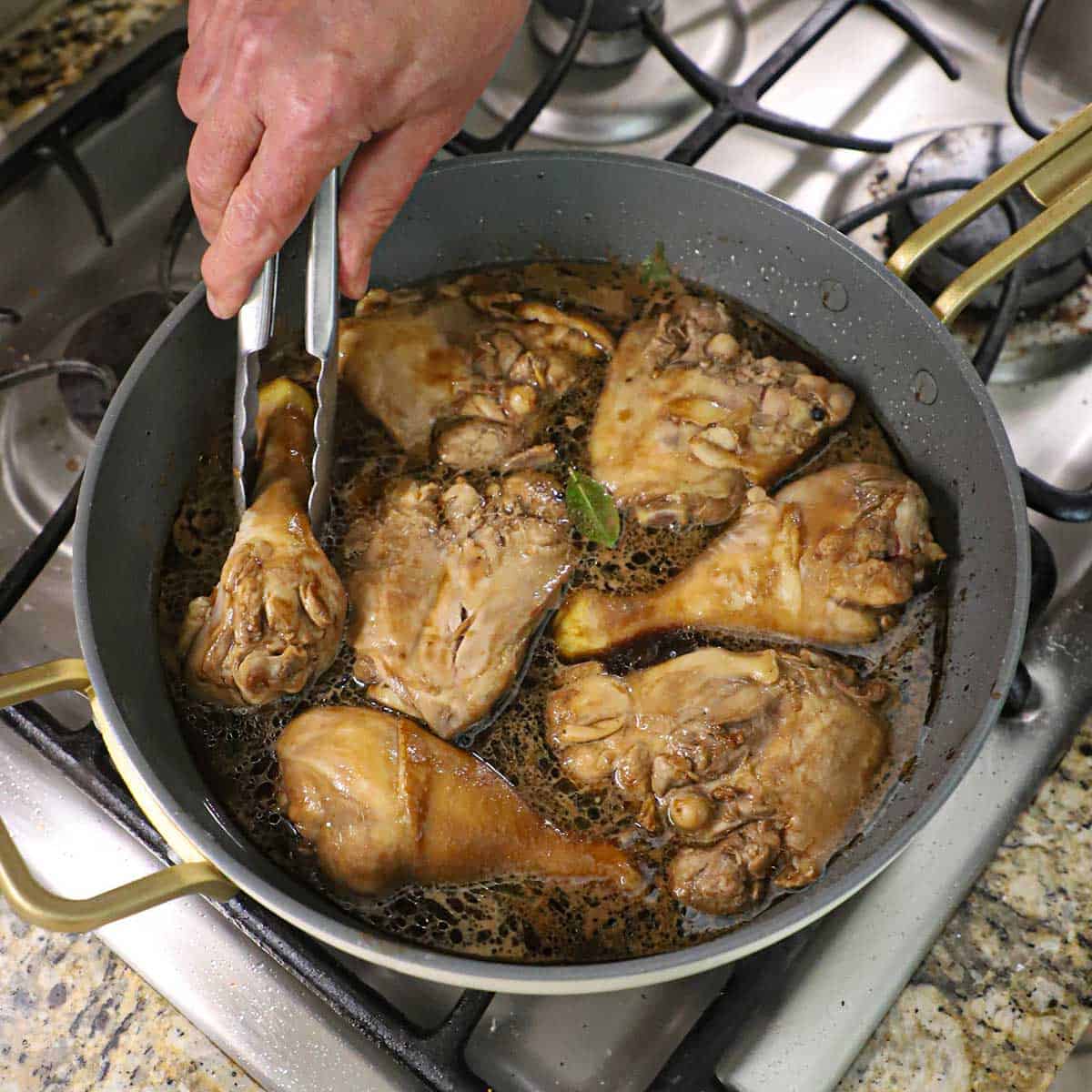 A person using a metal pair of tongs to place seared marinated chicken pieces into a skillet filled with a simmering Filipino chicken adobo sauce.