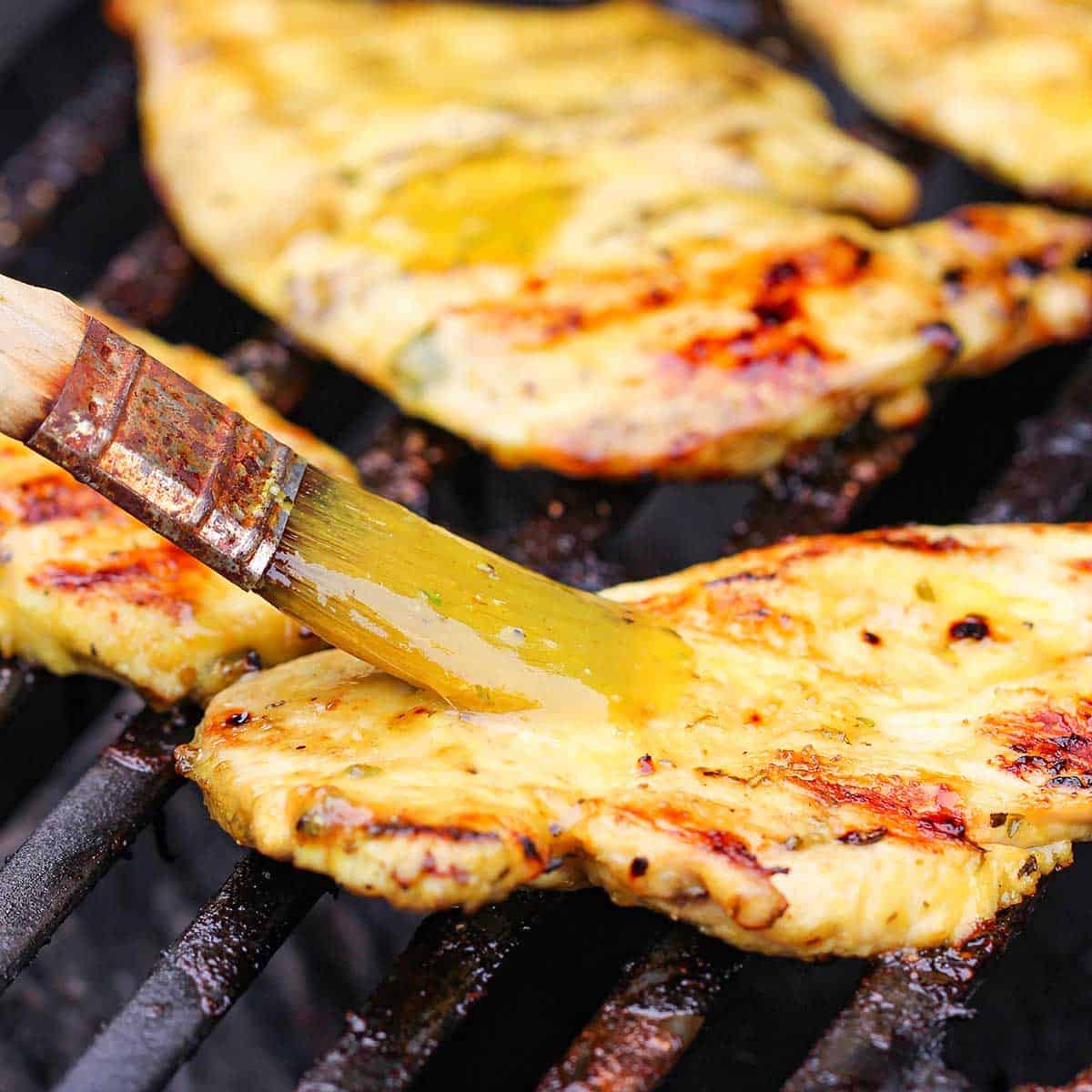 A person using a pastry brush to apply a honey mustard sauce onto a thin boneless chicken breast that is being grilled on a gas grill.