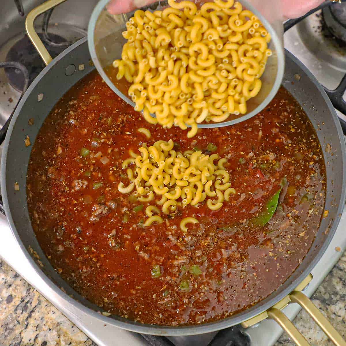 A person dumping uncooked small elbow macaroni from a glass bowl into a skillet filled with a red simmering American-style goulash meat sauce.