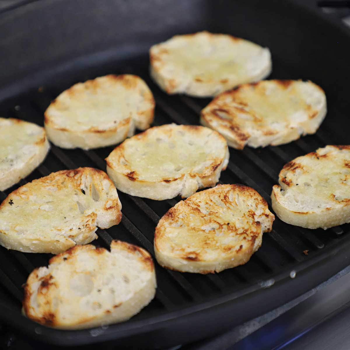 Thin slices of a baguette that are being toasted on a grill pan on a gas stove.