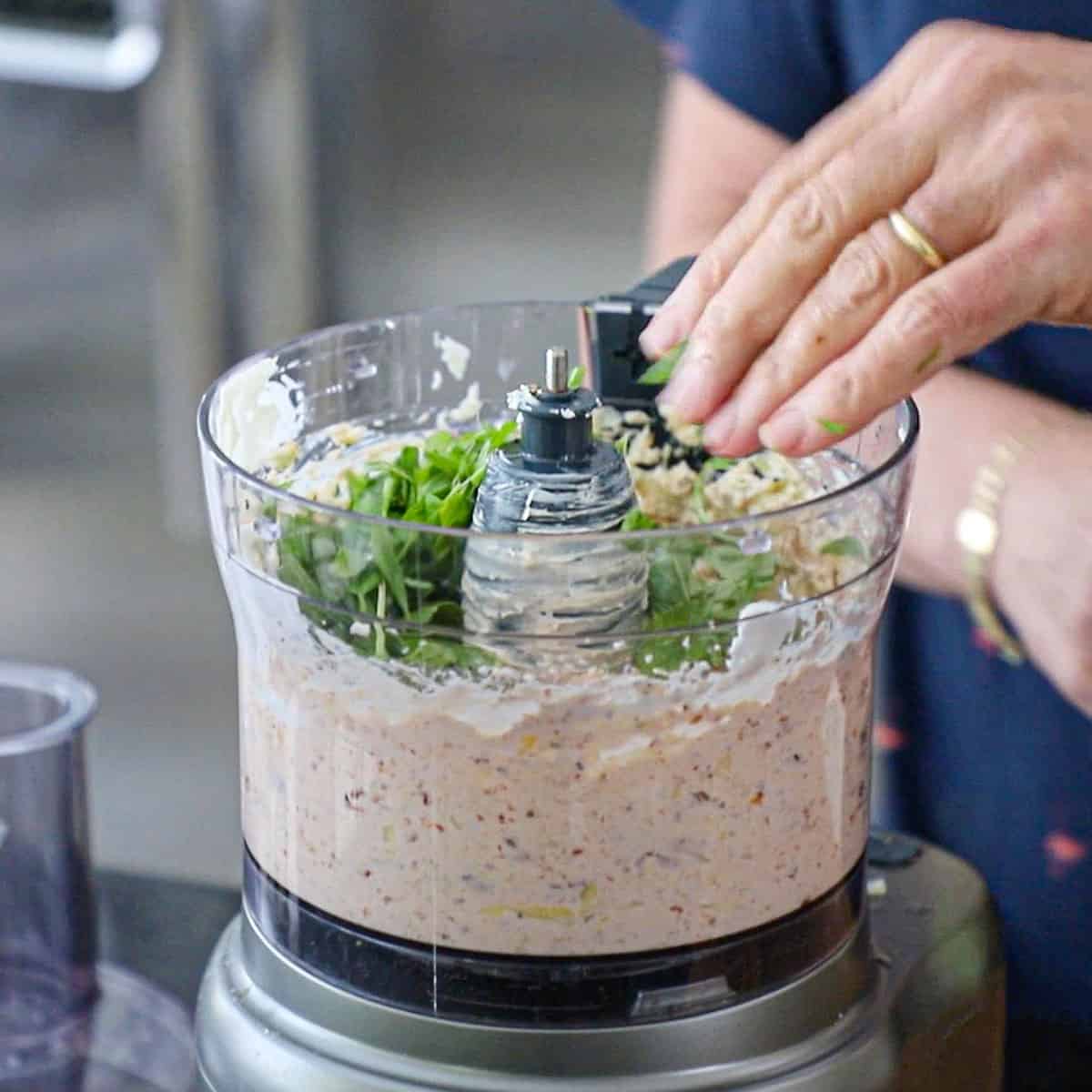 A person adding freshly chopped basil into a food processor that is filled with a smooth sun-dried tomato and artichoke dip mixture.