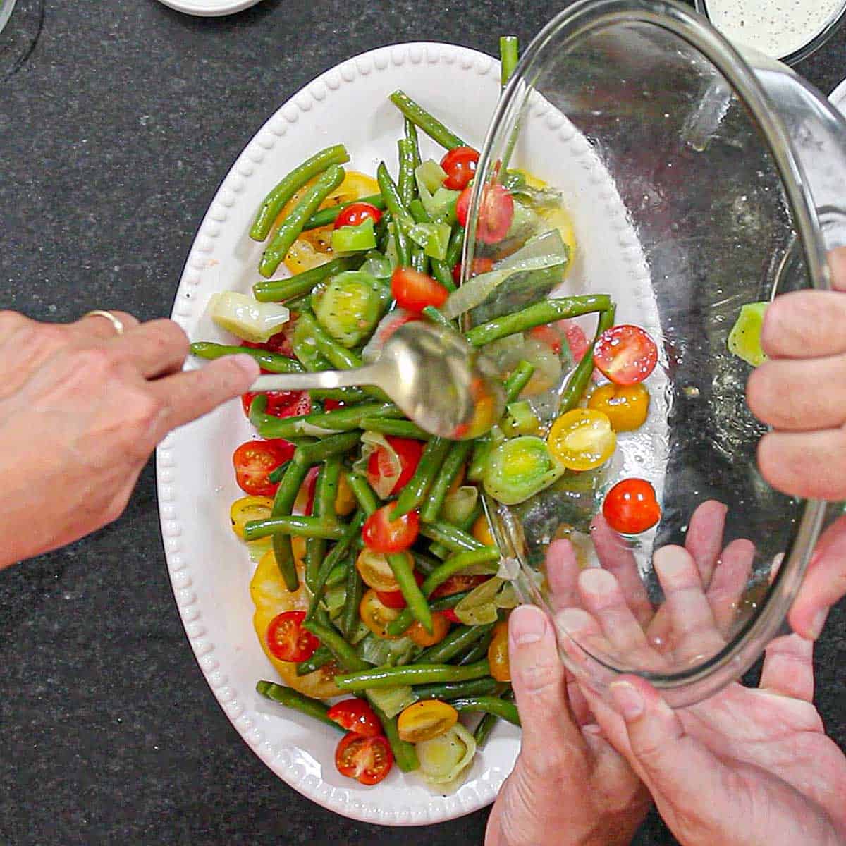 A person transferring a mixture of simmered green beans and leeks, combined with seasoned cherry tomato onto a white platter with sliced heirloom tomatoes on it.