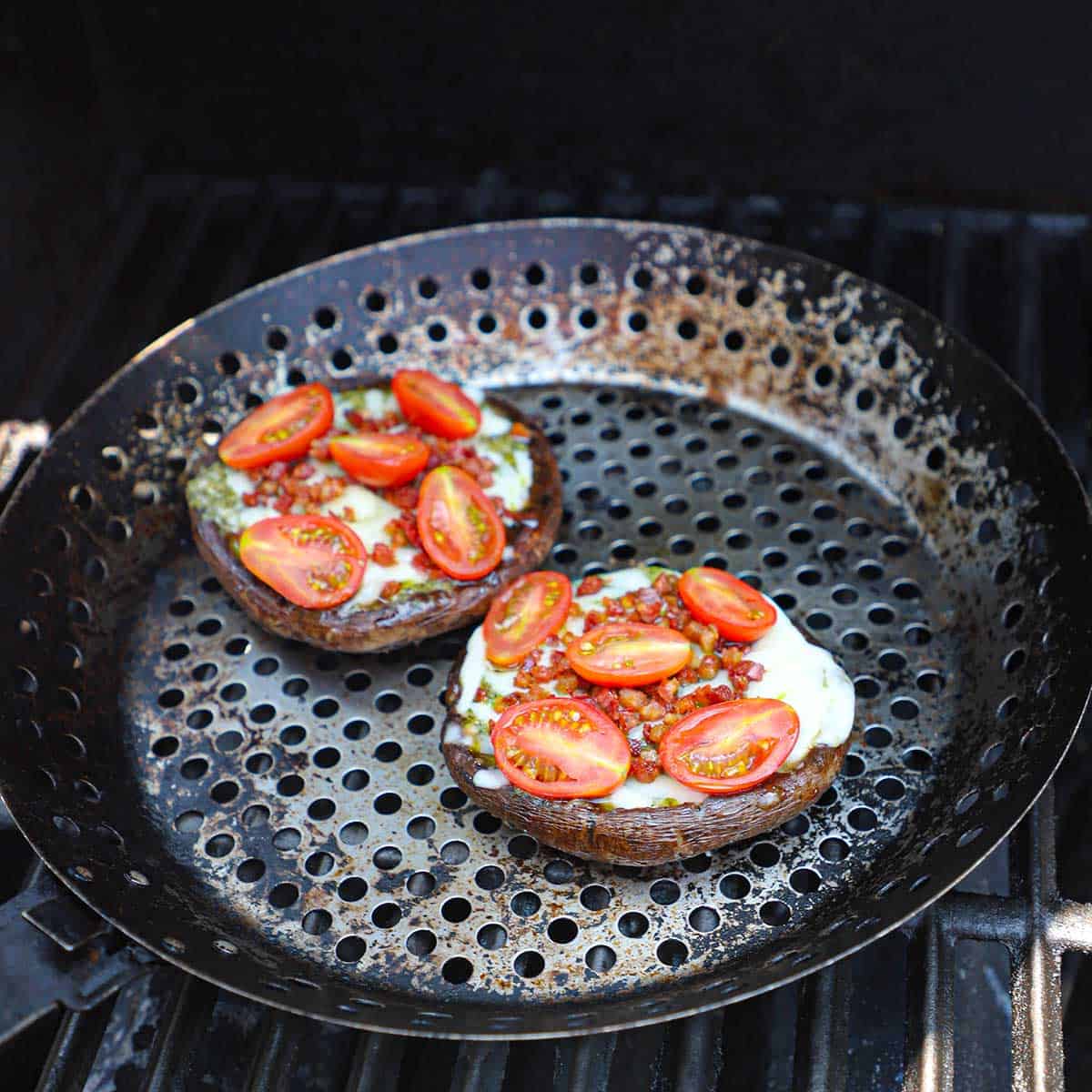 Two large portobello mushroom caps with cheese, pancetta, and cherry tomatoes being grilled in a circular grill pan on a gas grill.