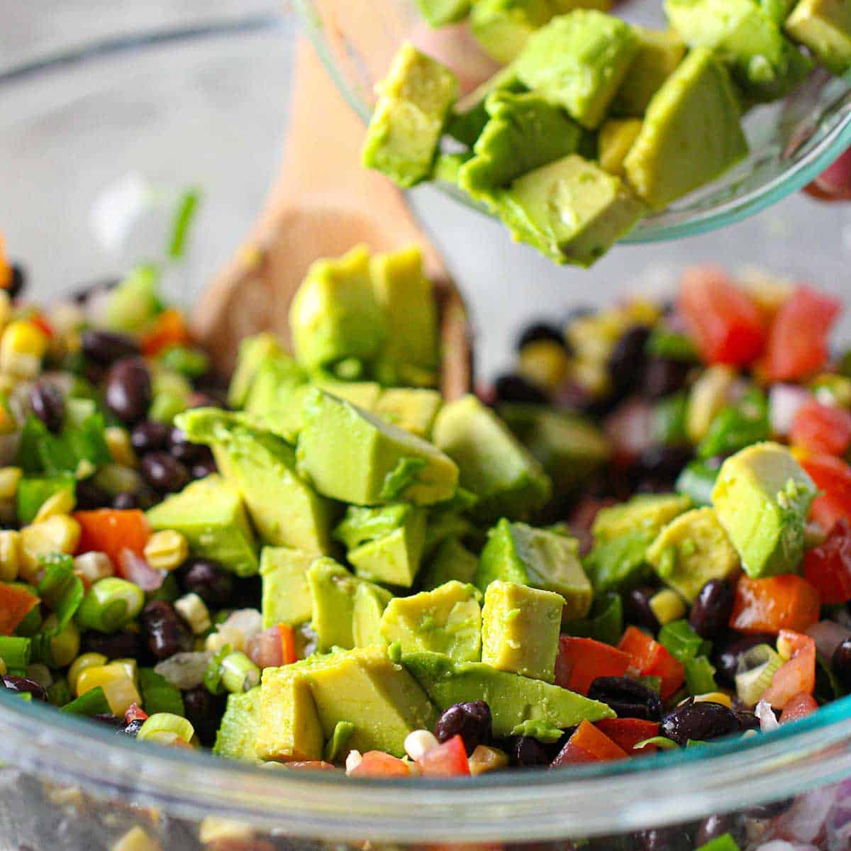 A person transferring cubed pieces of fresh avocado into a glass bowl of black beans, chopped tomatoes, and diced red onion.