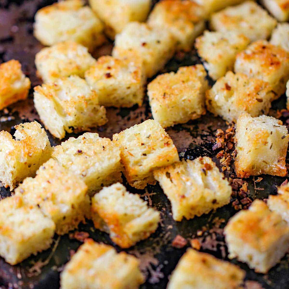 Freshly toasted homemade croutons that have been lightly seasoned and are resting on a scratched metal black baking pan.