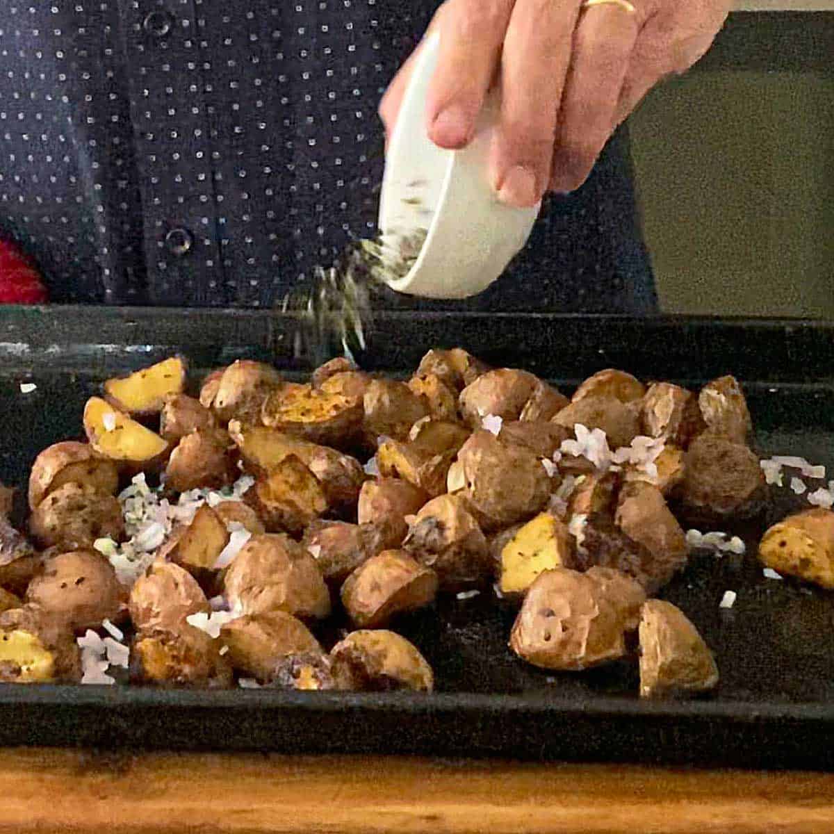A person tossing fresh herbs onto new potatoes that have been partially roasted on a baking sheet.