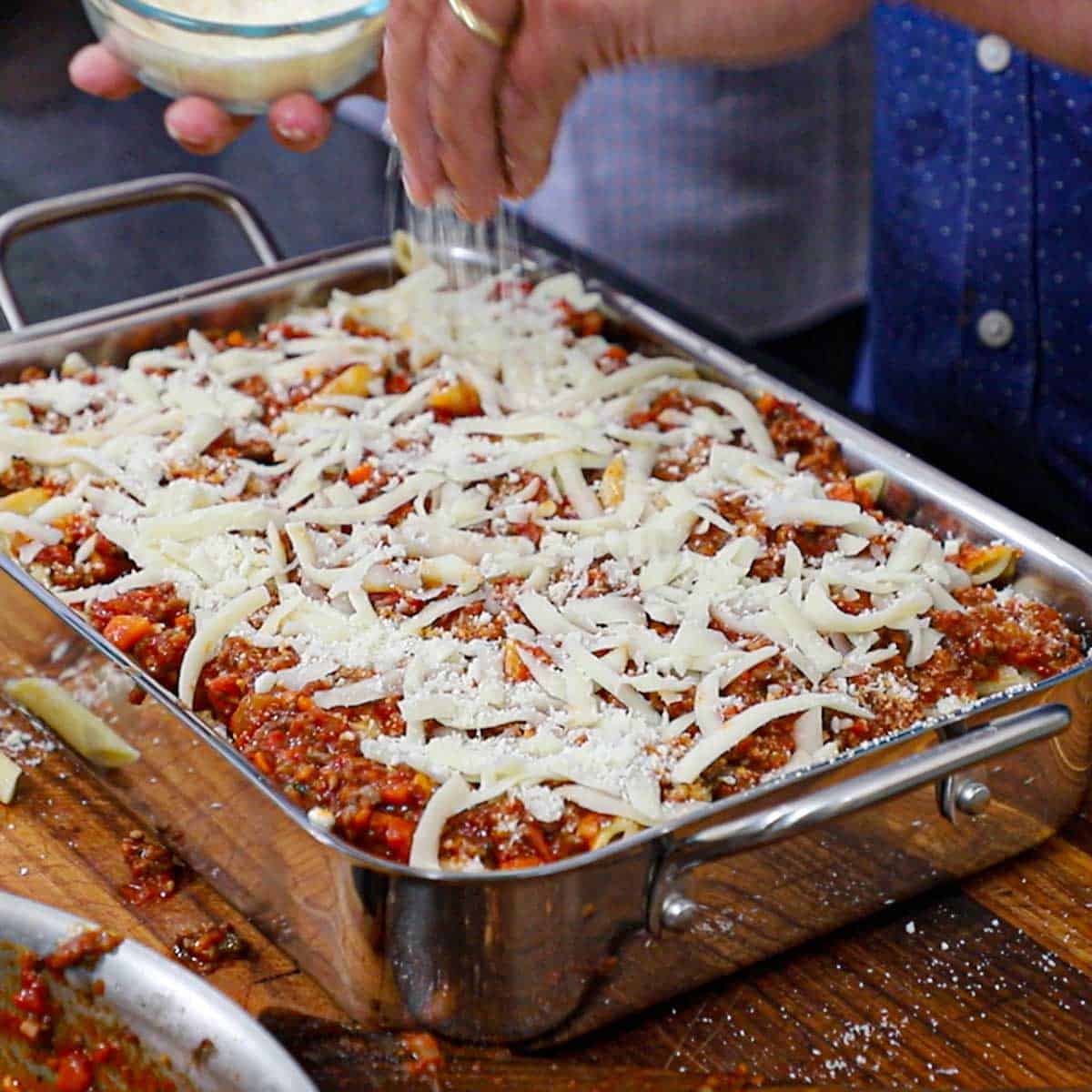 A person sprinkling grated parmesan cheese over the top of a silver lasagna pan that is filled with an uncooked baked ziti.