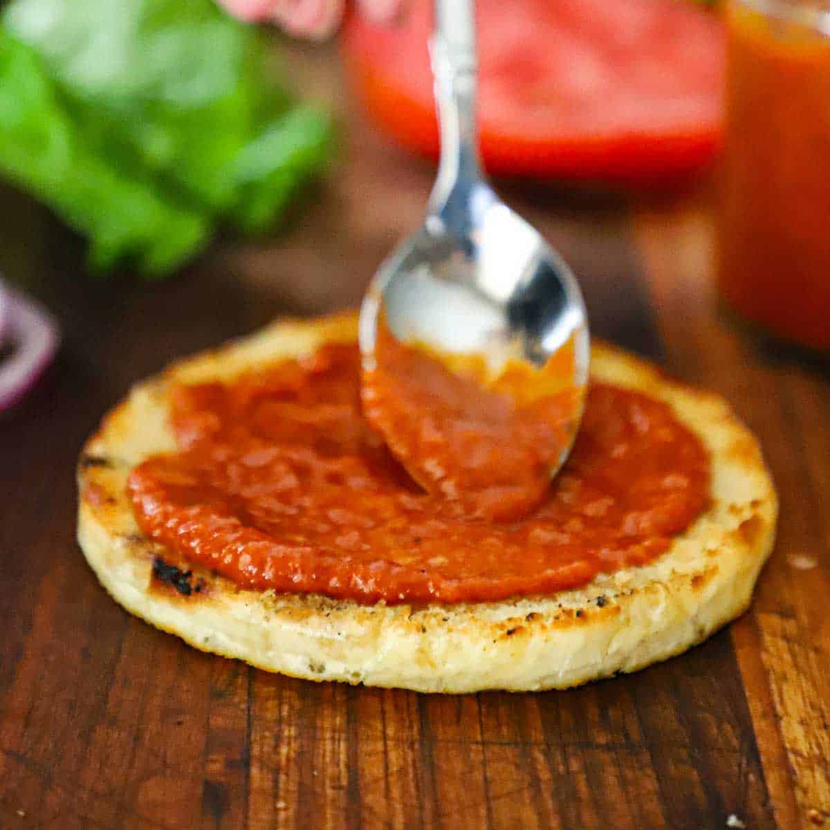 A person using a spoon to spread homemade ketchup on a bottom half of a toasted homemade hamburger bun resting on a wooden cutting board.
