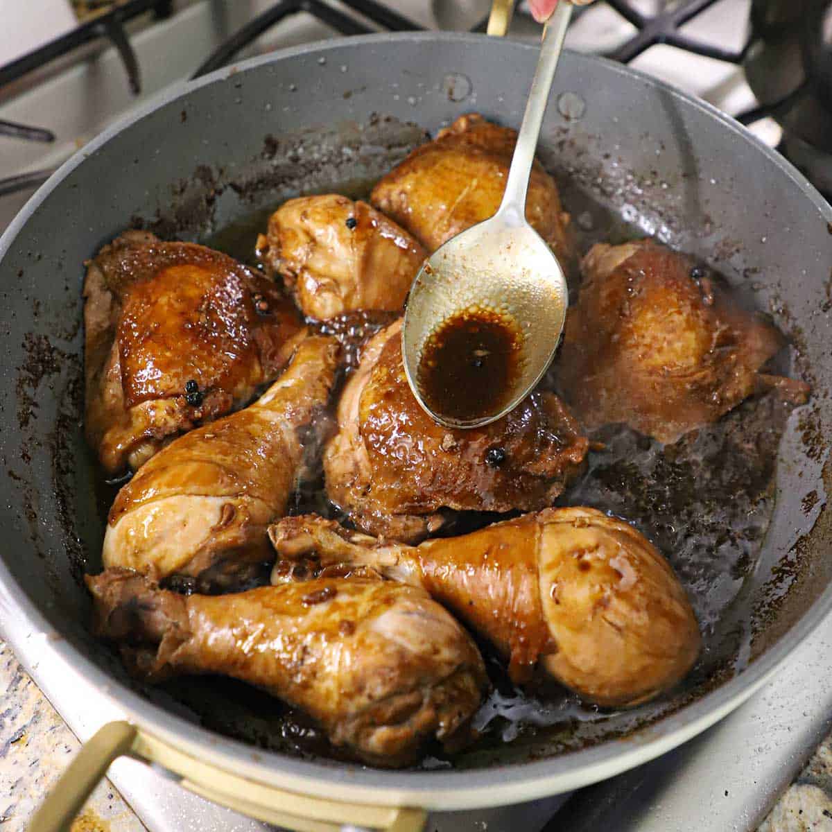 A person using a large spoon to basted chicken pieces that are simmering in a Filipino chicken adobo sauce on a gas stove.