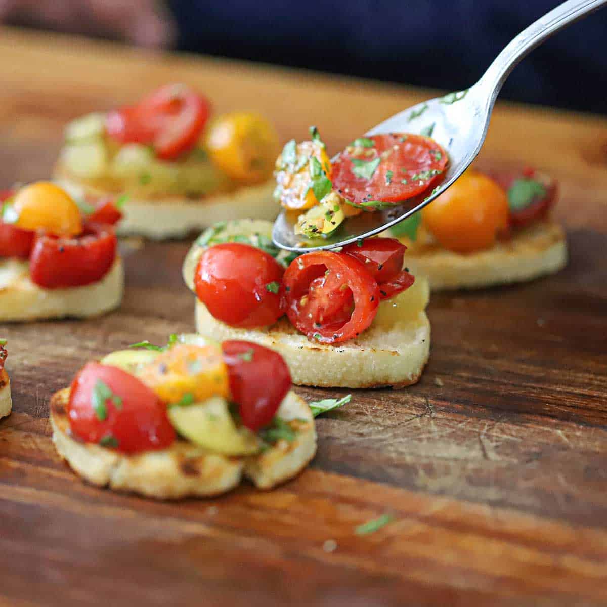 A person using a spoon to transfer a cherry tomato and basil topping onto a toasted slice of a baguette with other Summer bruschetta with tomato and basil sitting nearby on a cutting board.