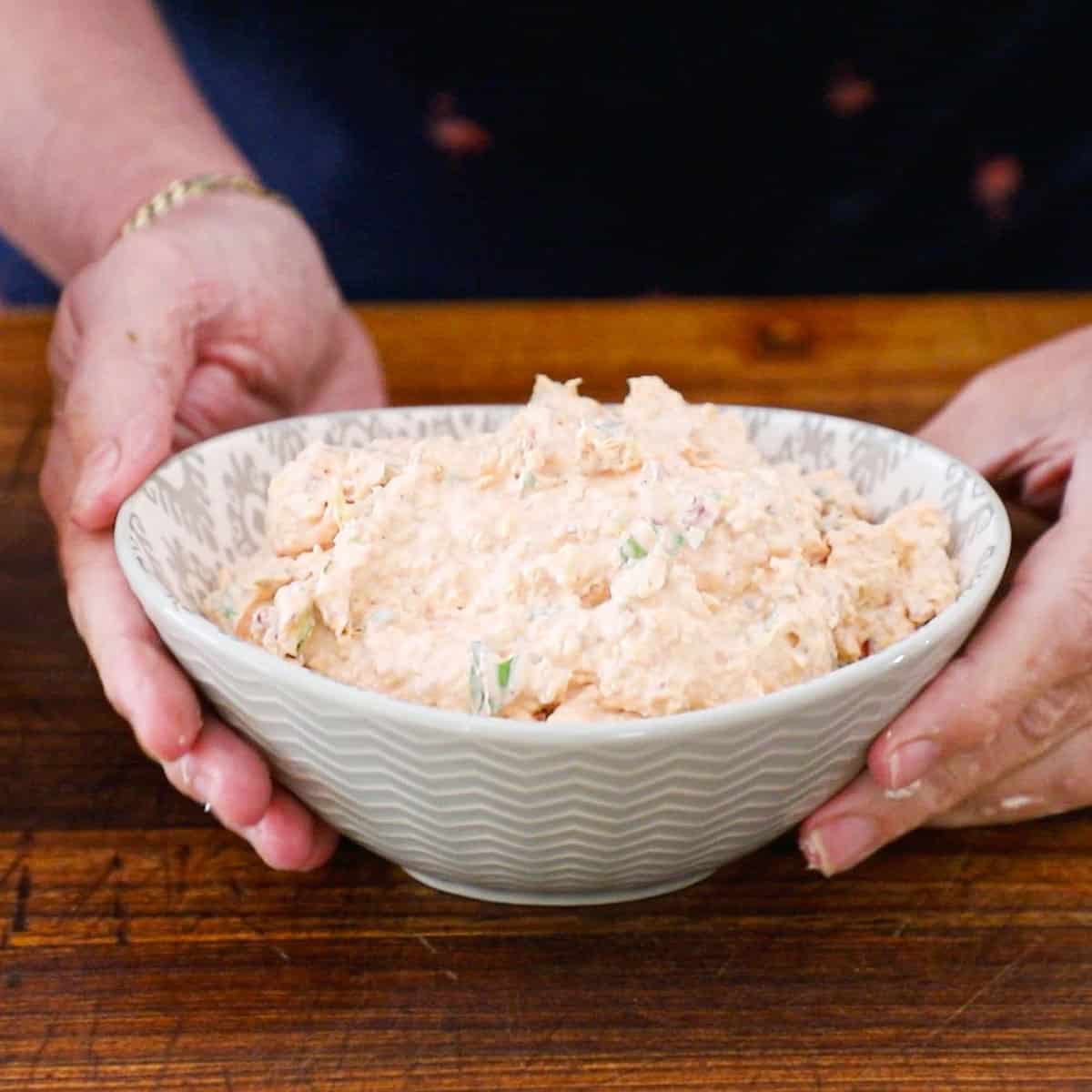 A person holding an oval bowl filled with sun-dried and artichoke dip over a wooden cutting board.