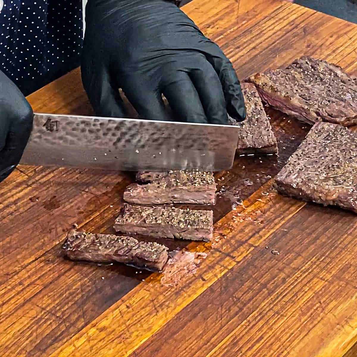 A person with black latex gloves using a large Japanese chef's knife to cut a grilled skirt steak into bite-sized slices on a wooden cutting board.
