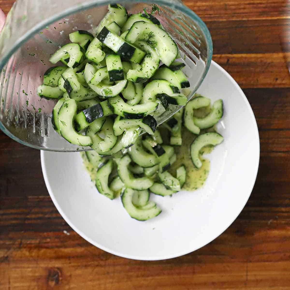 A person transferring a freshly prepared cucumber and dill salad from a glass bowl into a white serving bowl.