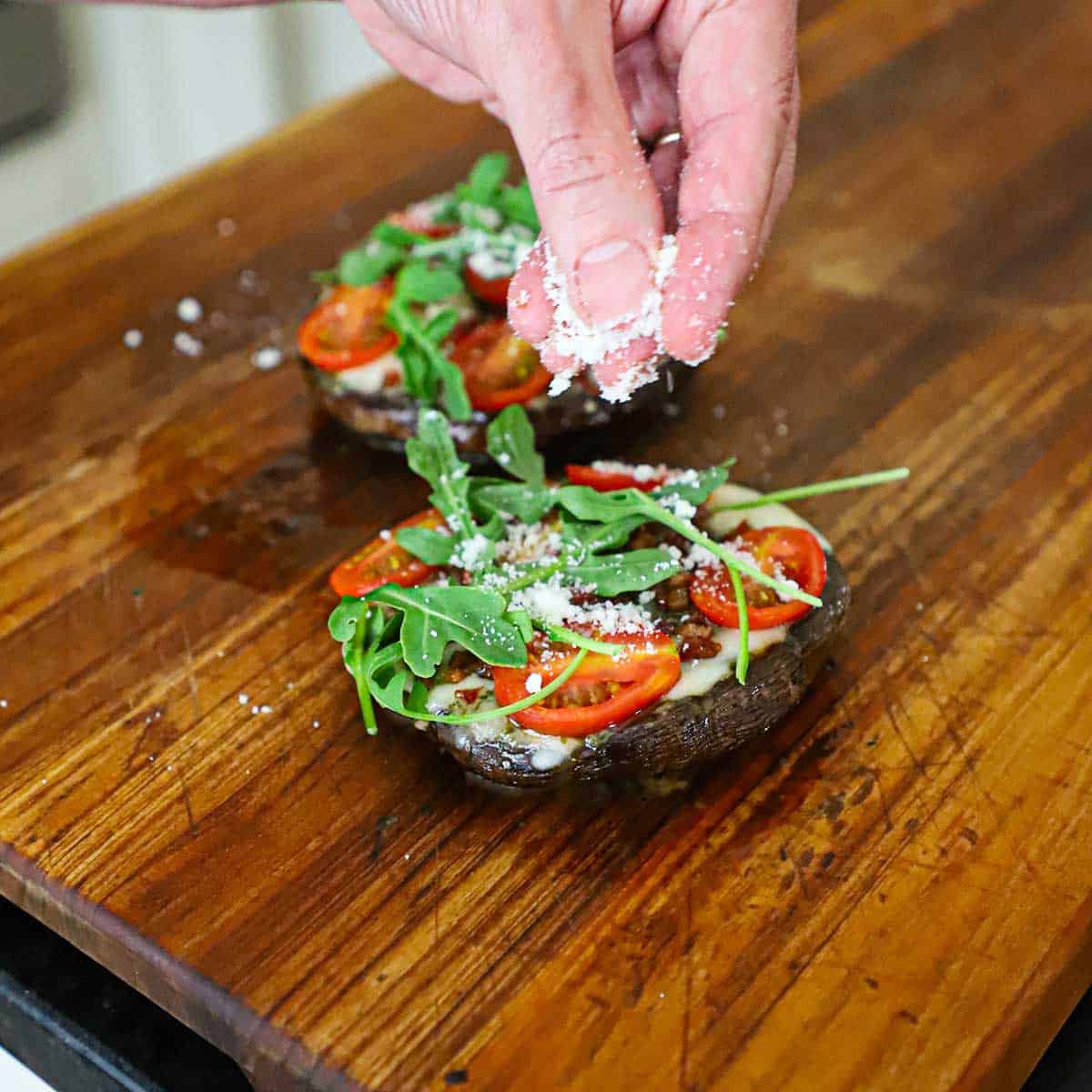 A person using his fingers to sprinkle grated Parmesan cheese over the top of a grilled portobello BLT pizza that is topped with sliced cherry tomatoes and arugula all on melted cheese and crispy pancetta.