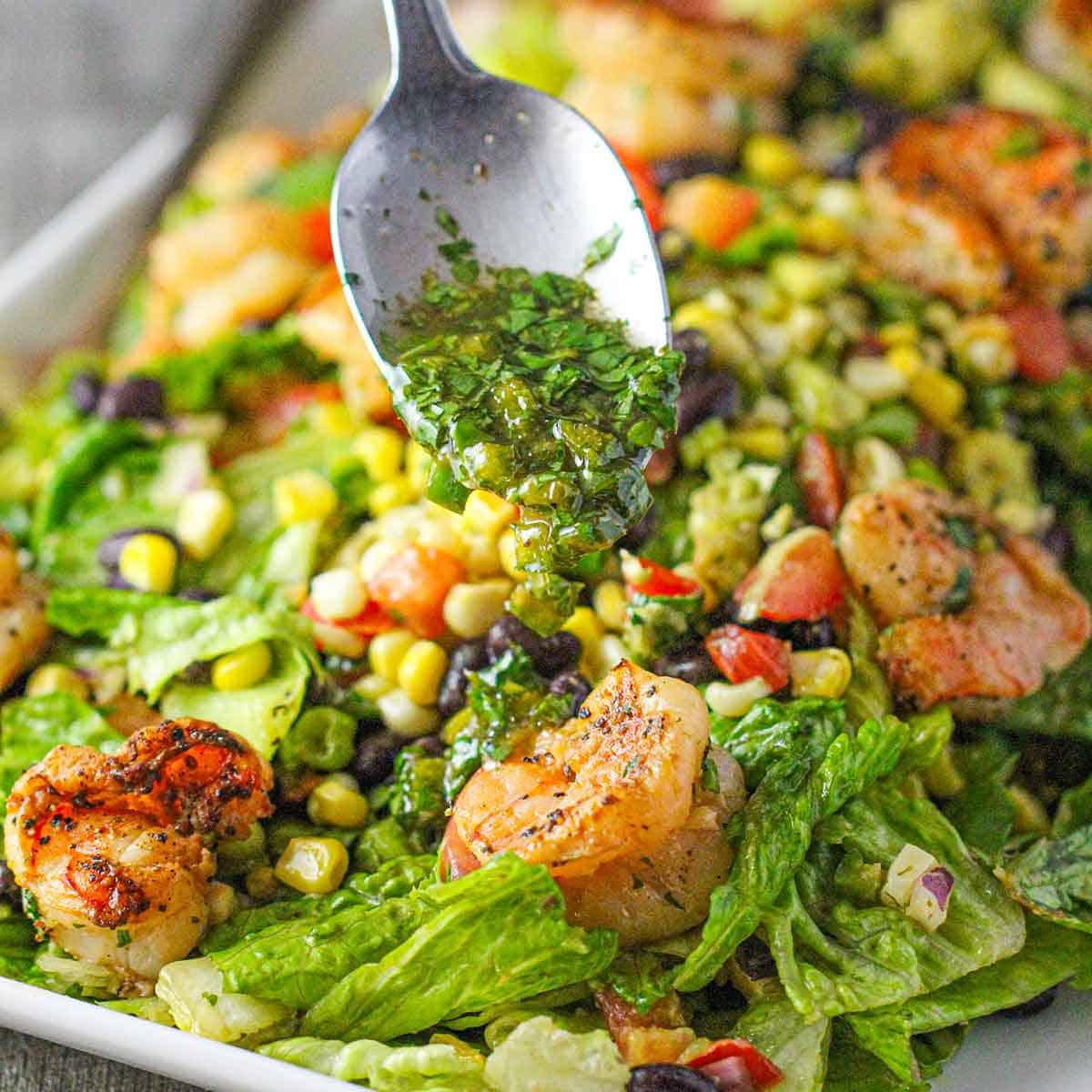 A person using a spoon to pour a cilantro-lime dressing over a salad of grilled shrimp, corn, black beans, tomatoes, and green leaf lettuce.