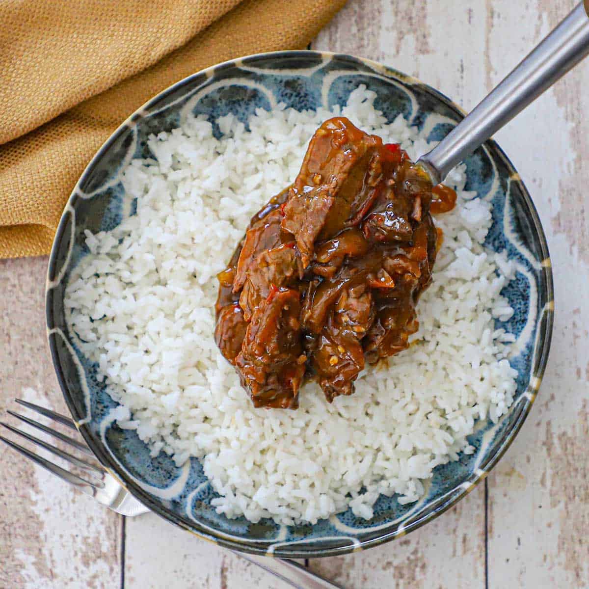 A person using a large serving spoon to place a mound of slow cooker pepper steak onto a shallow bowl filled with steamed white rice.