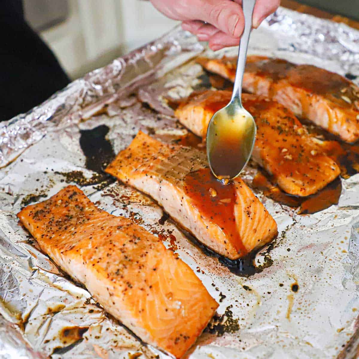 A person spooning a teriyaki glaze over freshly baked marinated salmon fillets that are resting on a foil-lined baking sheet.