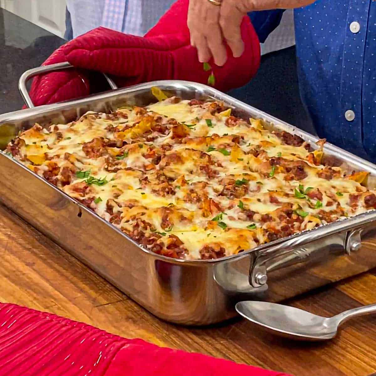 A person sprinkling freshly chopped Italian parsley over the top of a freshly baked ziti casserole in a silver lasagna pan resting on a wooden cutting board.