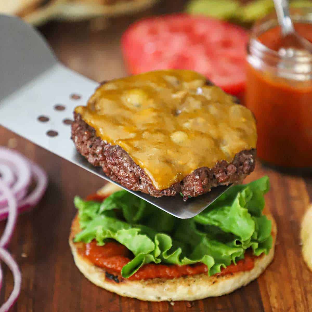 A person sliding a grilled hamburger patty with cheese onto the bottom half of a toasted hamburger bun that is topped with homemade ketchup and green leaf lettuce.