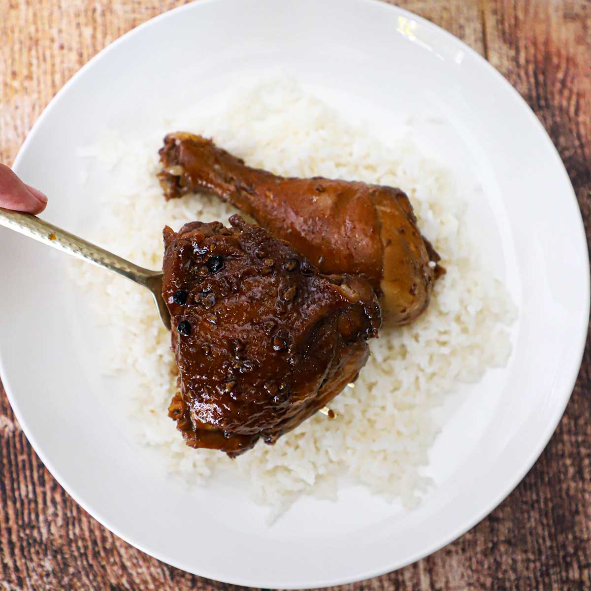 A person placing a fully cooked Filipino chicken adobo thigh onto a plate of steamed white rice and a cooked chicken leg.