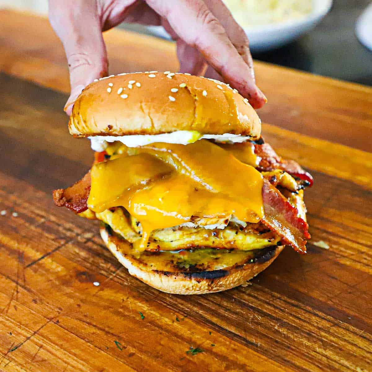 A person placing the top half of a toasted sesame seed bun on top of a grilled honey mustard sandwich with bacon on a cutting board.