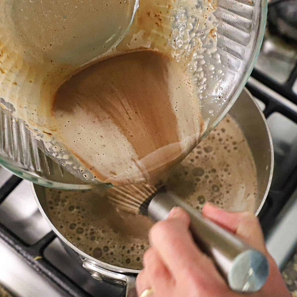 A person pouring a warm chocolate cream and egg mixture that has been tempered back into a saucepan on the stove.