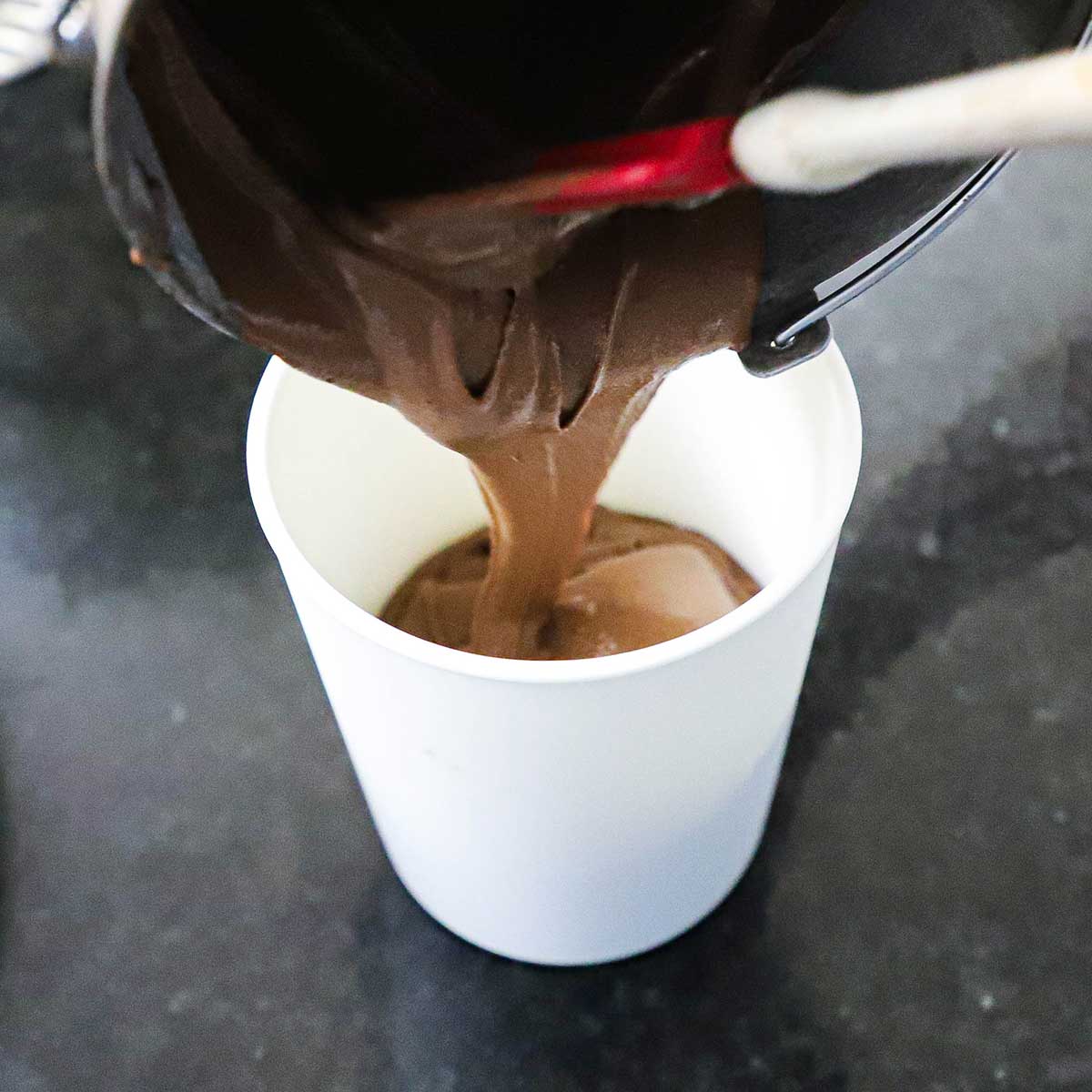 A person transferring freshly churned chocolate ice cream that is still soft into a white ice cream tub that will then go into the freezer.