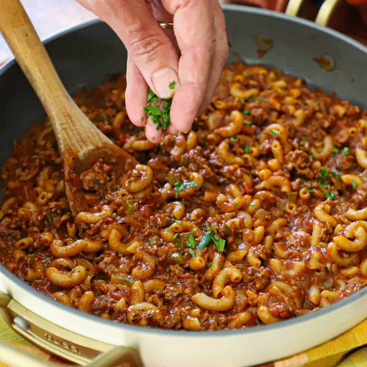 a person sprinkling chopped Italian parsley over the top of a skillet full of Mom's goulash (American-style) with a wooden spoon inserted into the middle of it.