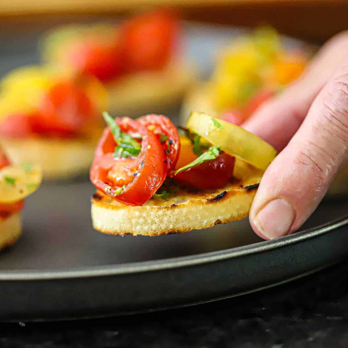 A person placing a Summer bruschetta with tomato and basil on a black plate next to more burshetta.