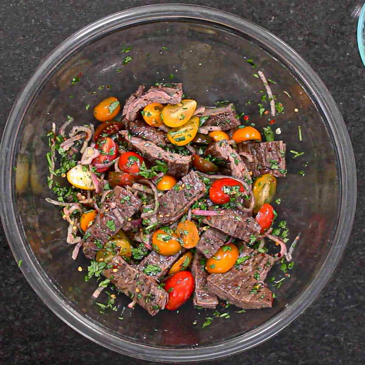 An overhead view of a glass bowl filled with strips of grilled skirt steak, halved cherry tomatoes, thin slices of shallot, herbs all tossed in a Thai sauce.