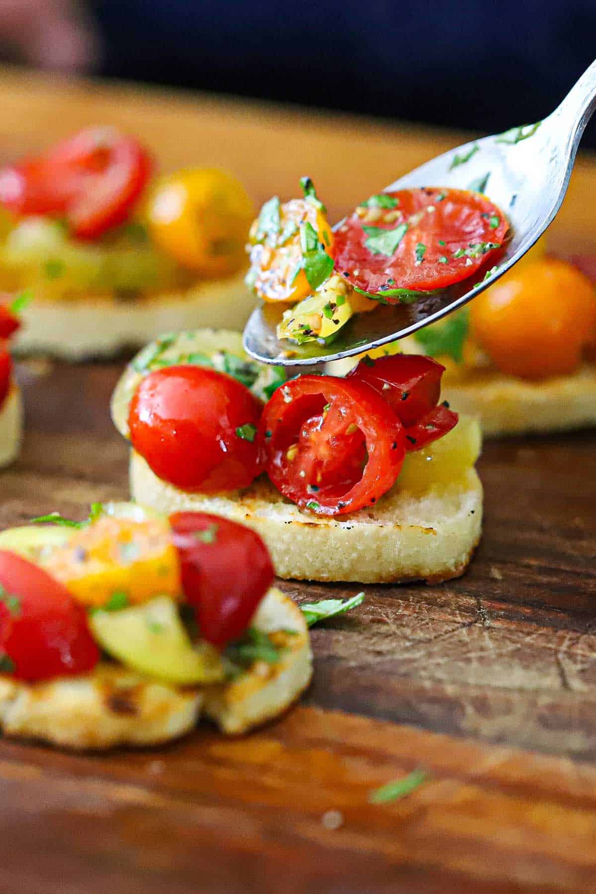 A person using a spoon to transfer a cherry tomato and basil topping onto a toasted slice of a baguette with other Summer bruschetta with tomato and basil sitting nearby on a cutting board.