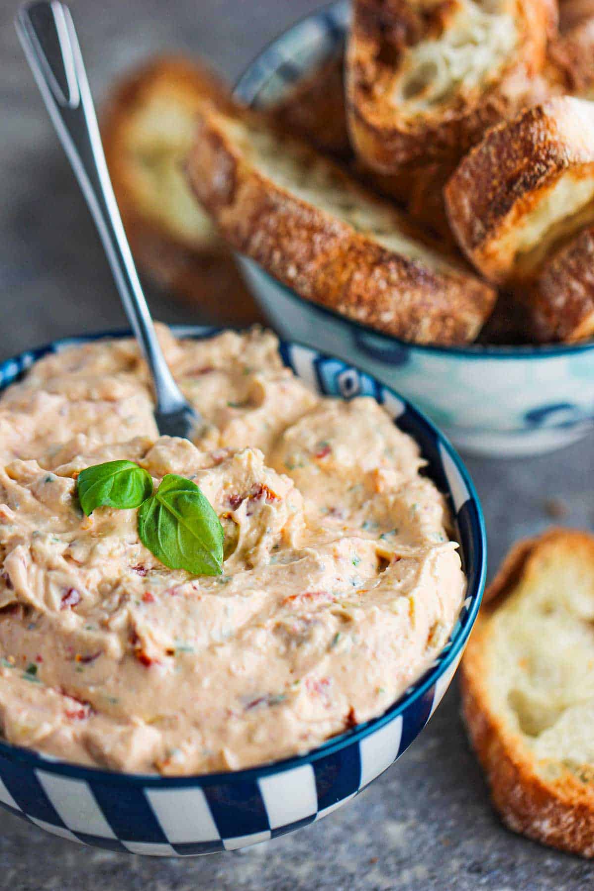 A colorful bowl filled with a sun-dried tomato and artichoke dip with another bowl filled with crusty pieces of sliced baguette.