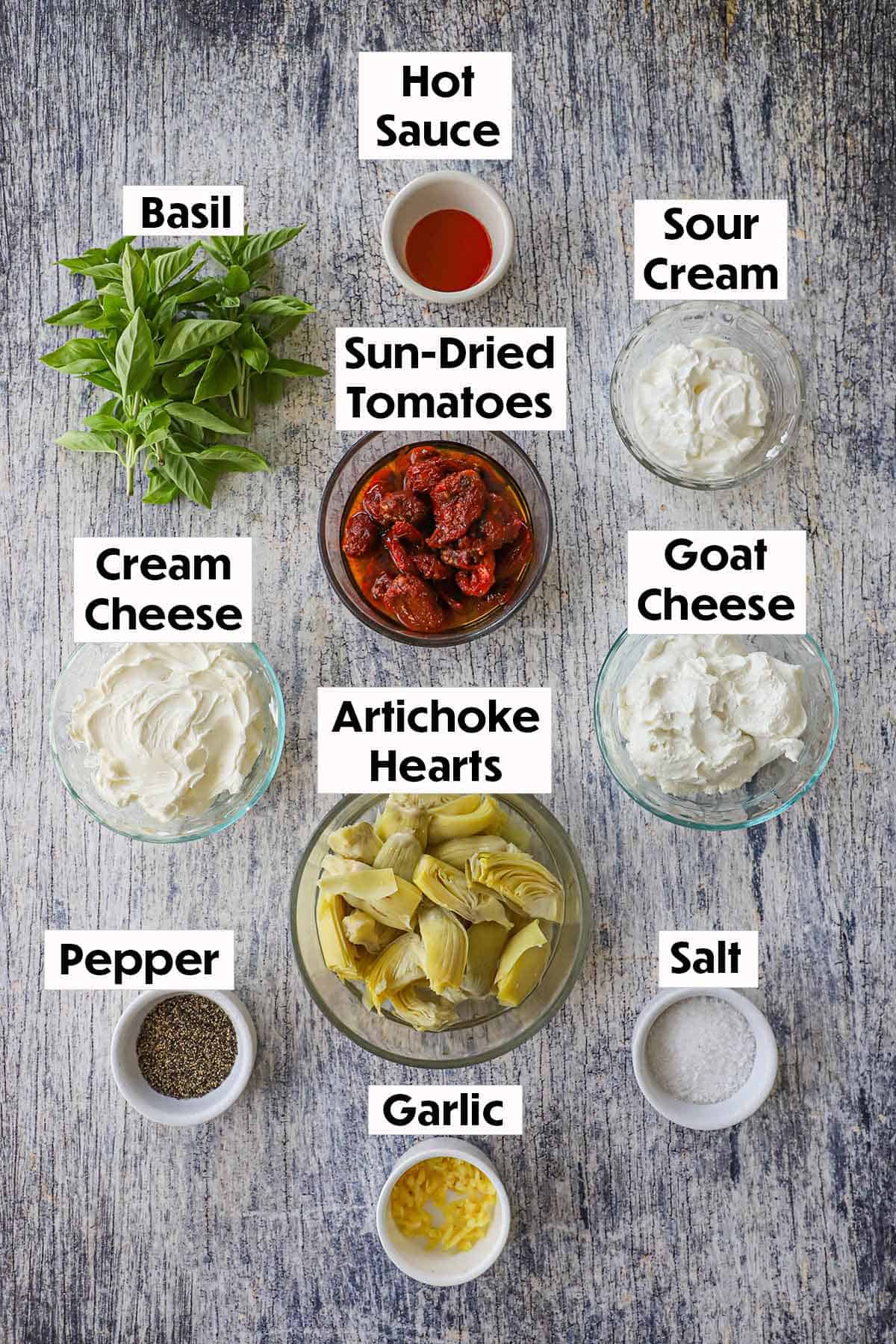 An arrangement of ingredients for sun-dried tomato and artichoke dip on a grey wooden background including sun-dried tomatoes, artichoke hearts, fresh basil, sour cream, goat cheese, cream cheese, and seasonings.