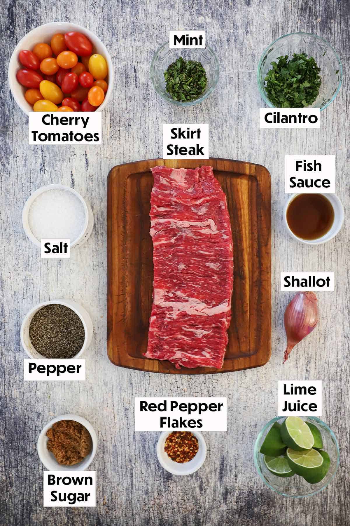 An arrangement of ingredients for Thai beef salad on a grey wooden background including an uncooked skirt steak, cherry tomatoes, herbs, seasonings, fish sauce, shallot, limes, and brown sugar.