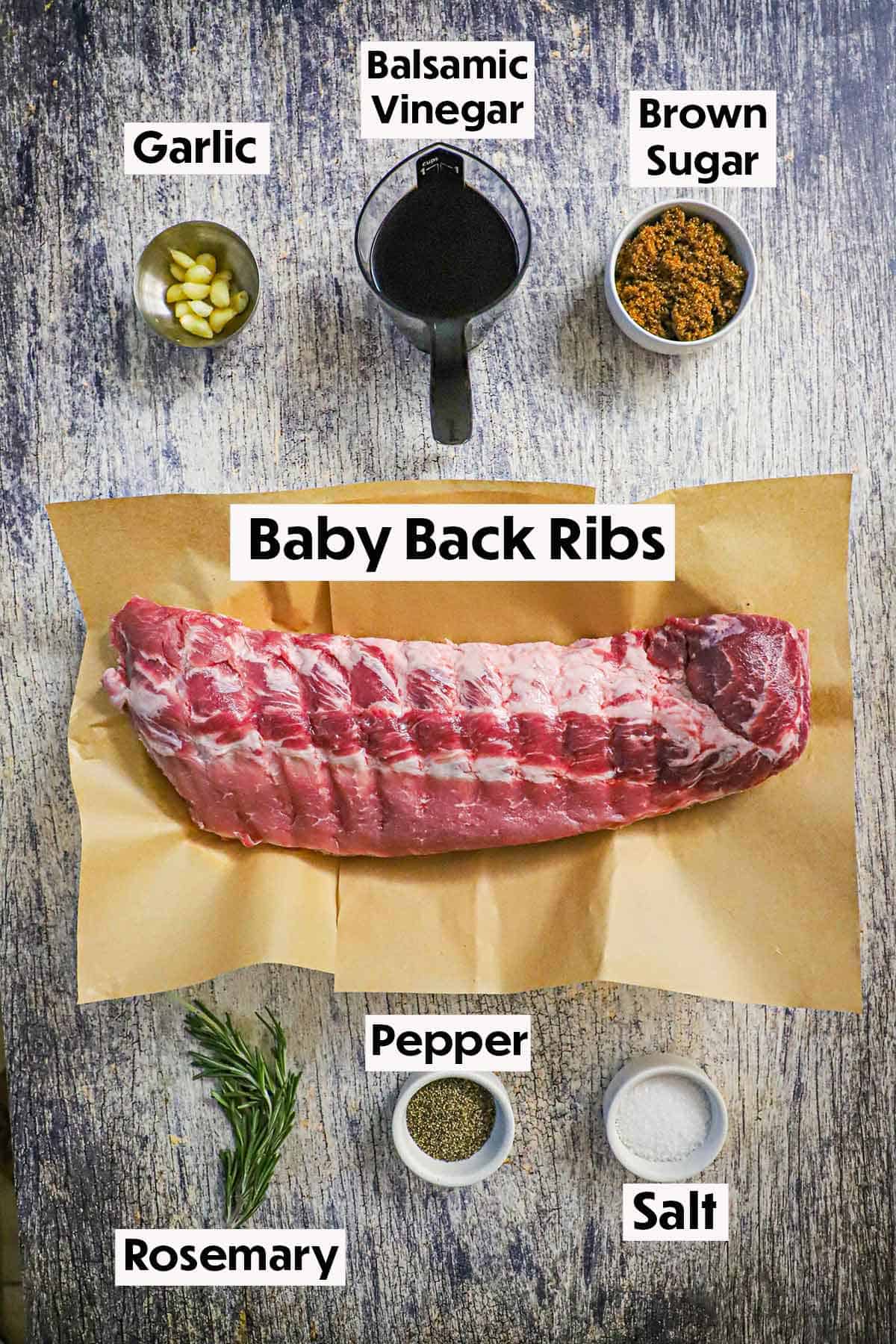 An arrangement of ingredients for balsamic-glazed baby back ribs on a wooden grey background including an uncooked rack of rbis, cloves of garlic, balsamic vinegar, brown sugar, fresh rosemary, salt, and pepper. 
