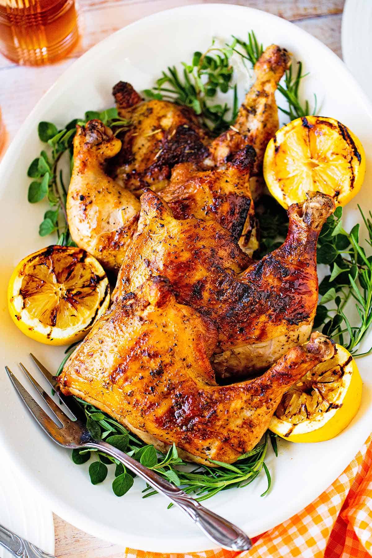 A large oval platter filled with four best grilled chicken quarters with fresh herbs and grilled lemon halves along the perimeter of the dish.