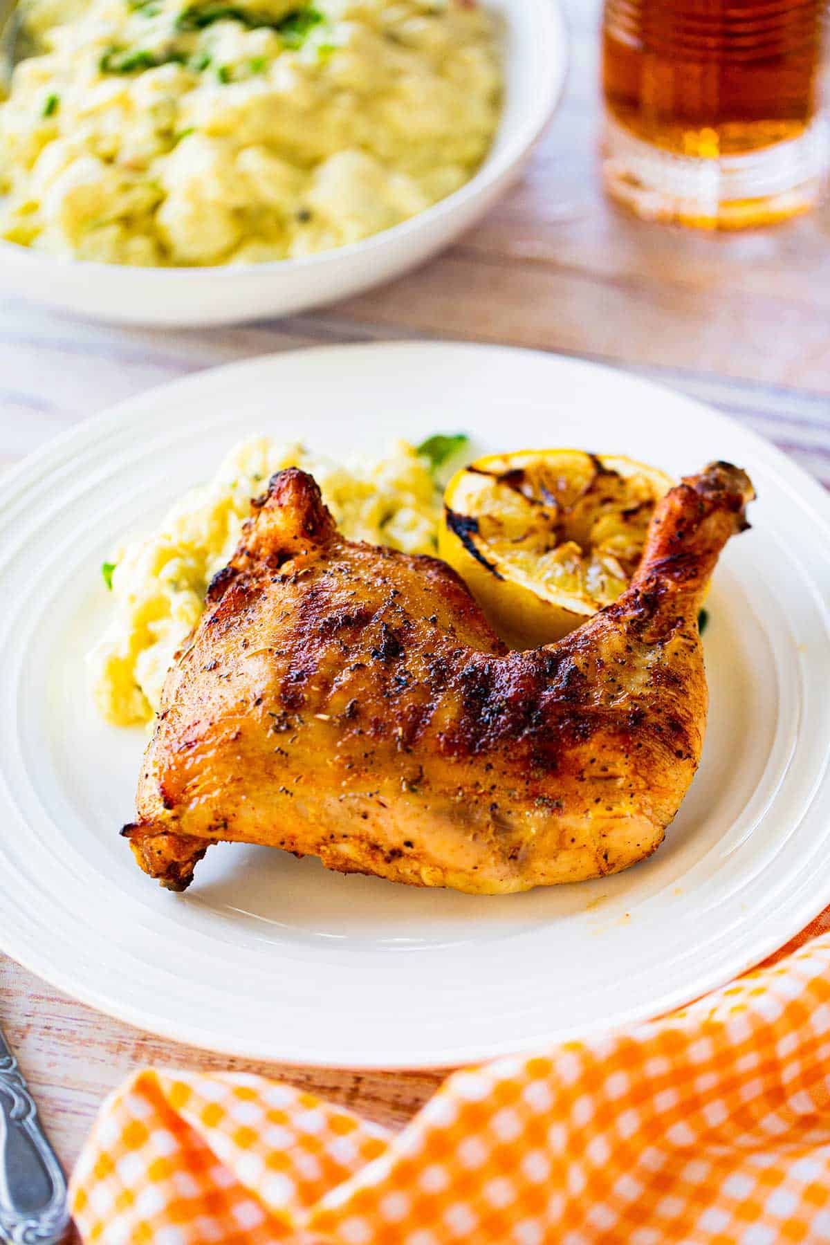 A best grilled chicken leg quarter resting on a white dinner plate next to a serving of potato salad and a grilled half of a lemon.