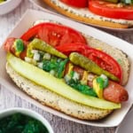 Two Chicago-style hot dogs in paper hot dog holders topped with a spear pickle, relish, peppers, and tomato wedges nestled around the hot dog inside a poppyseed bun.