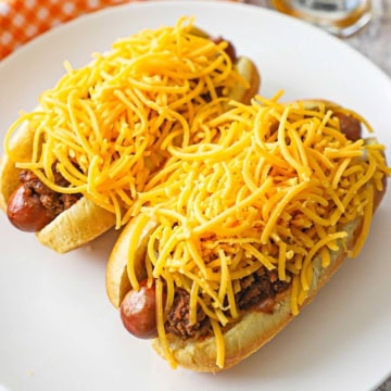 Two Cincinnati Cheese Coney hot dogs sitting on a white dinner plate, both dogs topped with large piles of shredded cheddar cheese.