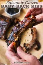 A person holding a balsamic glazed baby back rib over a trey of the entire rack of ribs with a small bowl of the glaze nearby.