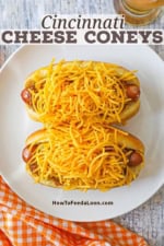 An overhead view of two Cincinnati cheese Coney hot dogs that are both piled high with shredded cheddar cheese on a white plate next to a tall glass of beer.