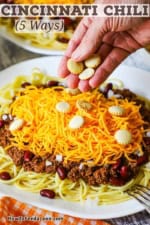 A person sprinkling oyster crackers over the top of a large serving of Cincinnati chili over spaghetti with a mound of shredded cheddar cheese on top.