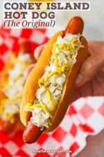 A person holding a Coney Island hot dog in his hand with the frankfurter nestled into the bun and topped with sauerkraut, chopped onions, and a zig zag of deli-style mustard.