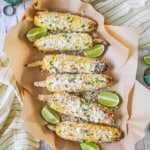 A row six ears of Mexican street corn, eloté, resting on sheets of brown paper with lime wedges scattered around them.