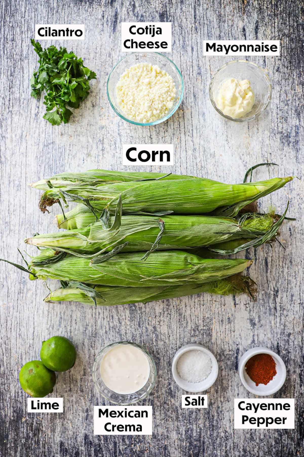 An arrangement of ingredients for eloté, Mexican street corn, on a wooden grey background including ears of corn in their husks, cilantro, cheese, mayonnaise, cream, seasonings, and limes.